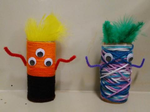 Two yarn monsters. One on the left made with orange and black yarn with two pipe cleaner arms, three googly eyes and yellow feathers. The one on the right made with variations of blue, purple, and white yarn with two pipe cleaner arms, two googly eyes, and green feathers.