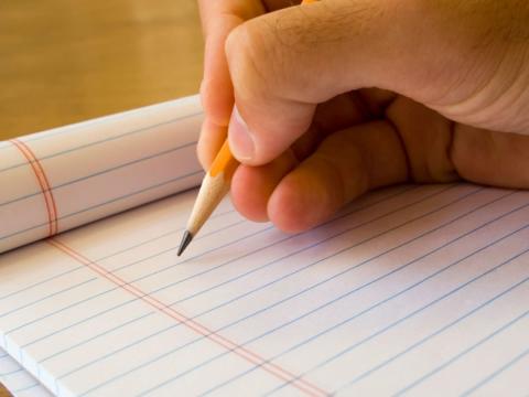 An image of a hand holding a yellow pencil above a pad of lined paper.